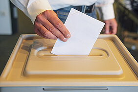Eine Hand steckt einen Zettel in eine Wahlurne ein