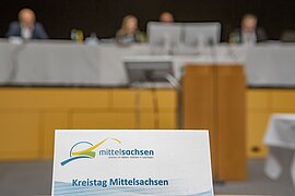 im Vordergrund ein Schild mit der Aufschrift Kreistag Mittelsachsen, im Hintergrund das Podium mit mehreren Verwaltungsangestellten