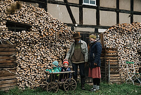 An einem Fachwerkhaus liegt ein Holzstapel, davor stehen ein Mann und eine Frau mit zwei Kindern, die in einem Handwagen sitzen.