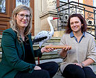 zwei Frauen sitzen auf einer Treppe und halten einen Staffelstab, im Hintergrund steht ein Deko-Storch