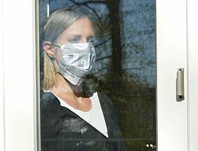 traurige Frau steht mit Mundschutz hinter Fenster