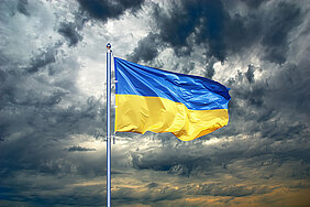 Dunkle Wolken am Himmel, davor weht die ukrainische Flagge