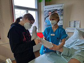 an einem Krankenbett unterstützt eine Krankenschwester eine Schülerin bei Überziehen eines medizinischen Strumpfes