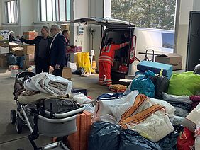 Zwei Männer stehen in einer Halle, die mit Säcken und Kisten gefüllt ist. Im Hintergrund wird ein Transporter mit Spenden beladen