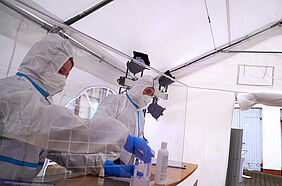 Zwei Mitarbeiter des Testzentrums stehen in Schutzkleidung hinter einer Glasscheibe. 