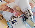 Eine Glühlampe liegt neben einem Netzstecker auf mehreren verschiedenen Geldscheinen
