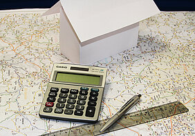 aus Papier gefaltetes Haus steht auf einer Landkarte, daneben liegen ein Stift und ein Lineal