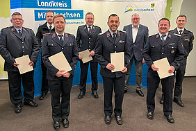Mehrere Feuerwehrleute in Uniform stehen mit Abstand vor einer Pressewand mit der Aufschrift Landkreis Mittelsachsen